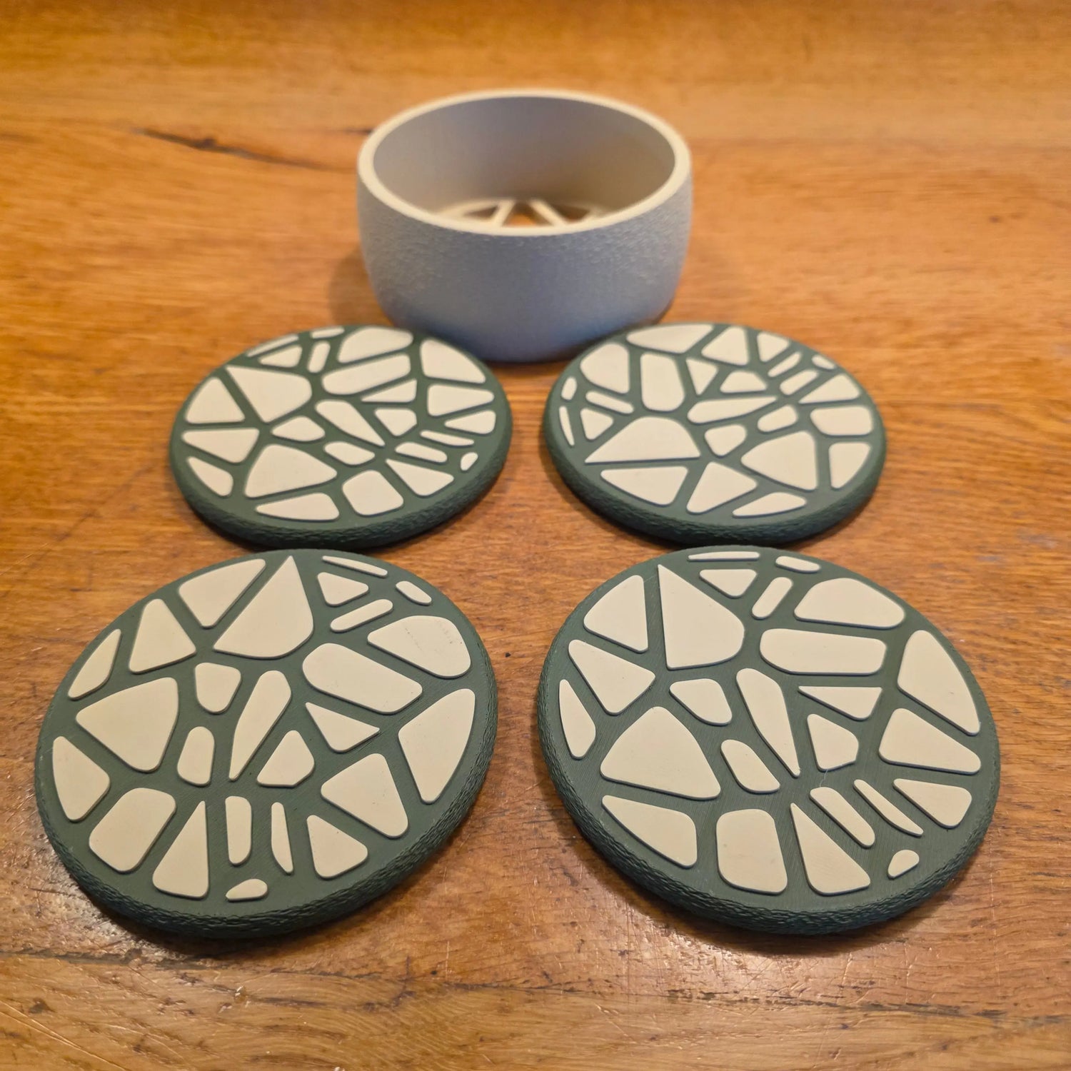 Ensemble de quatre dessous de verre au design Voronoi géométrique, imprimés en 3D en plastique noir mat, disposés sur une table en bois clair.